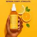 Hand holding Skynfluent Sacred Citrus Scalp Tonic with fresh orange and lemon slices – scalp spray to refresh, purify, and stimulate.
